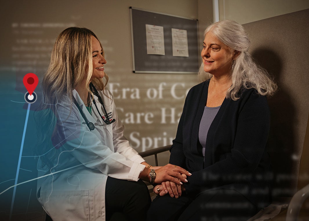 Dr. Gloria Ethier holds an elderly, silver-haired white woman's hands as they sit and smile during a patient consultation.