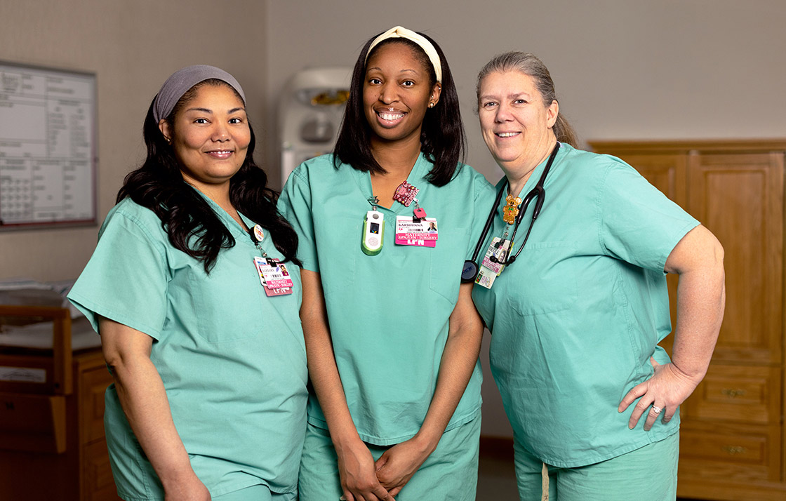Three female nurses of diverse ethnicities and ages stand together in the specialized baby nursery within the maternity ward.