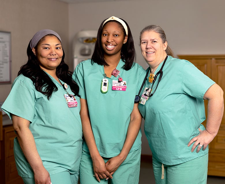 Three female nurses of diverse ethnicities and ages stand together in the specialized baby nursery within the maternity ward.