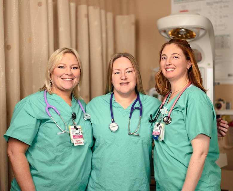 A trio of neonatal nurses wearing green scrubs and stethoscopes stands smiling in the Burdett Specialized Care Nursery.